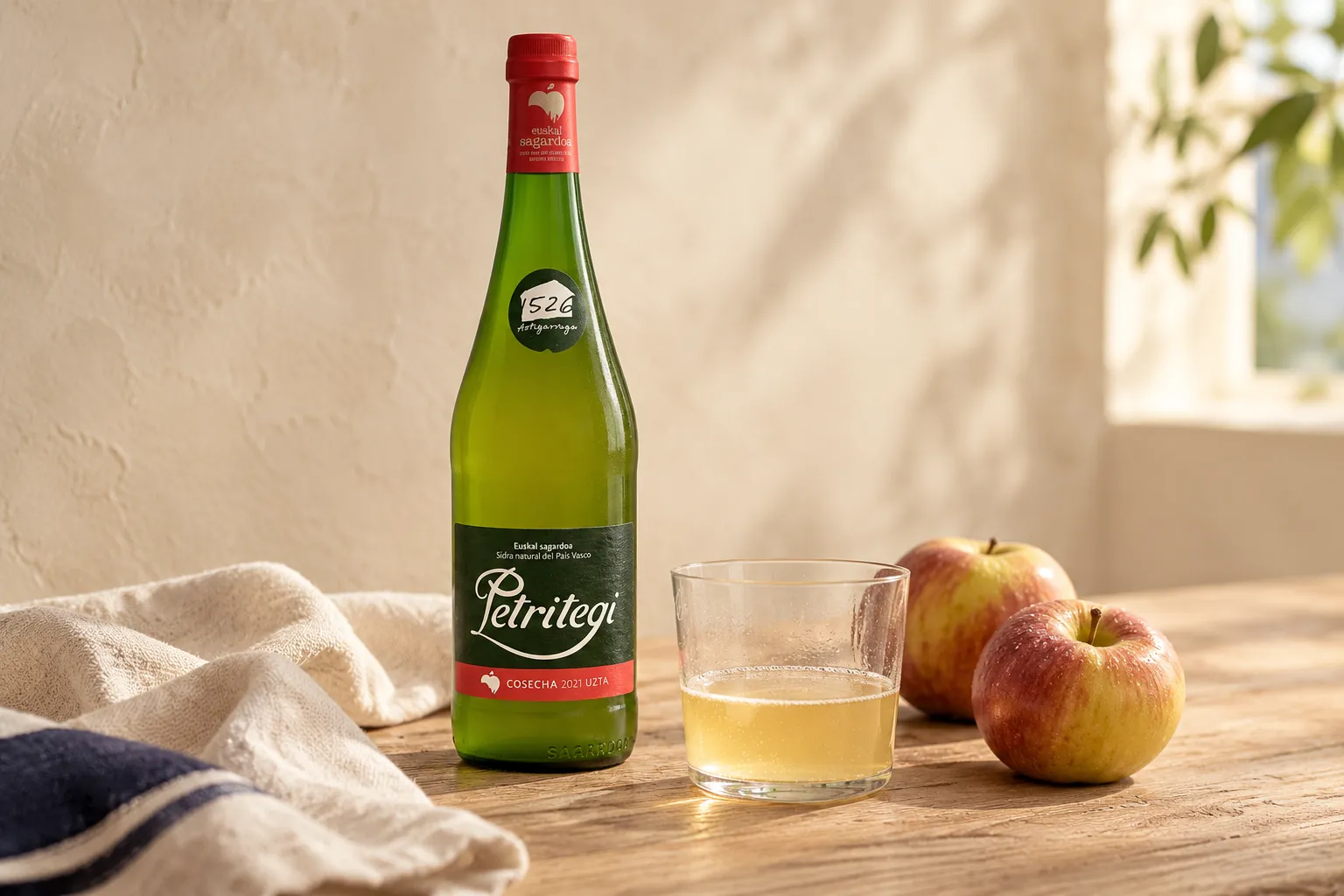 Botella de sidra Petritegi junto a un vaso servido y dos manzanas sobre una mesa de madera.