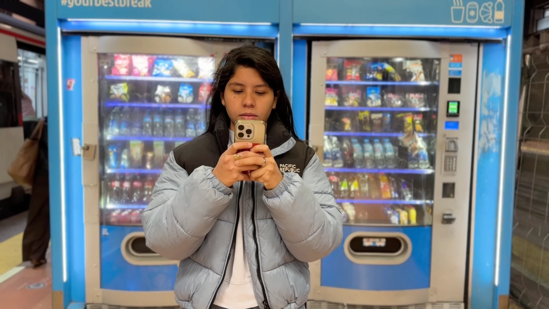 Zabrinna mira su móvil de pie frente a dos máquinas expendedoras en un andén de metro.